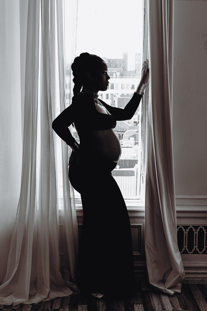 black and white pregnancy photo of an African American woman standing at a window in profile by Leona Darnell.