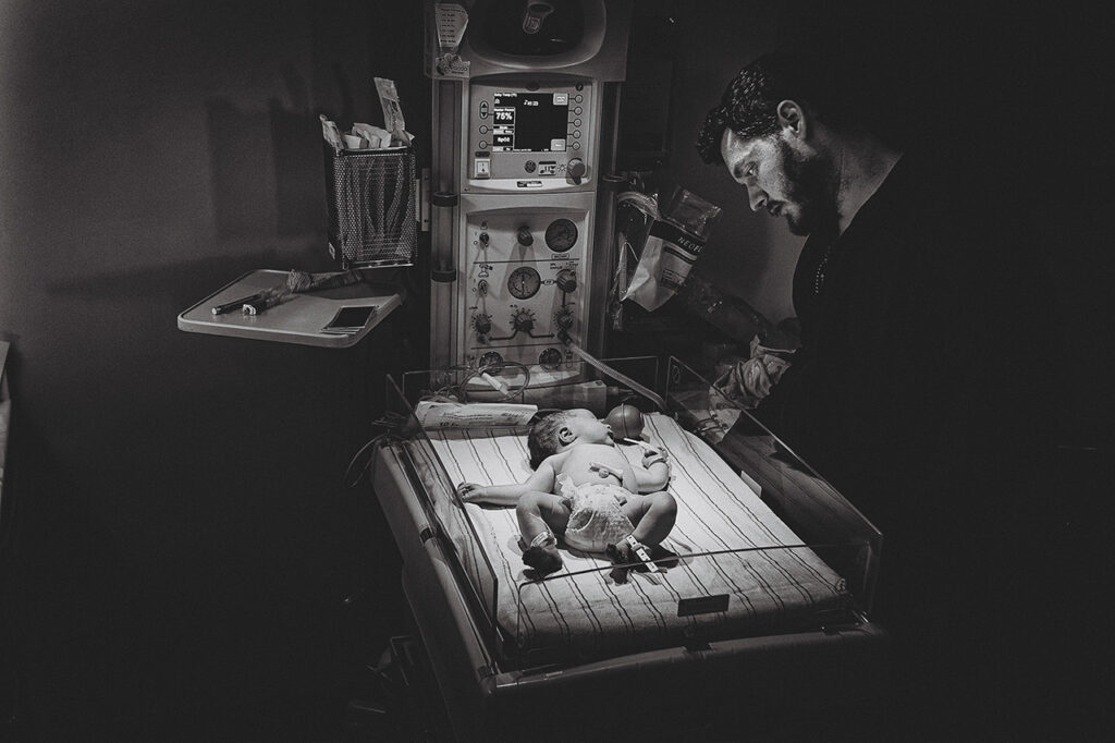 Black and white birth photography photo of a man looking at his infant son in the warmer by Leona Darnell.