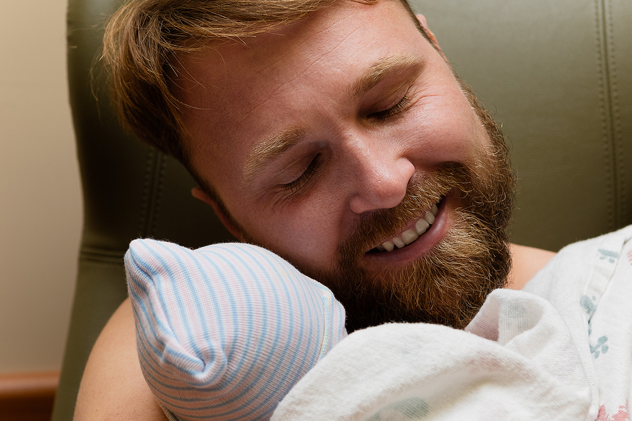 A father smiling while holding his newborn daughter by birth photographer, Leona Darnell