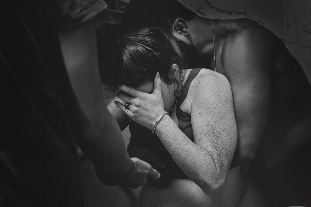 black and white image by Los Angeles doula and birth photographer, Leona Darnell. This shows a woman being supported by her husband and doula,